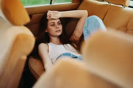 Расслабленная молодая женщина сидит в винтажном автомобиле, одетая в белую майку и синие джинсы, с задумчивым видом на фоне тёплого, землистого интерьера, который вызывает чувства ностальгии и…