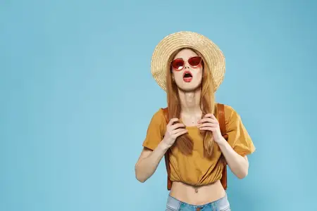 Красивая женщина в шляпе, темных очках и модной одежде, демонстрирующая эмоции в студии, кадрированный вид, веселая модель