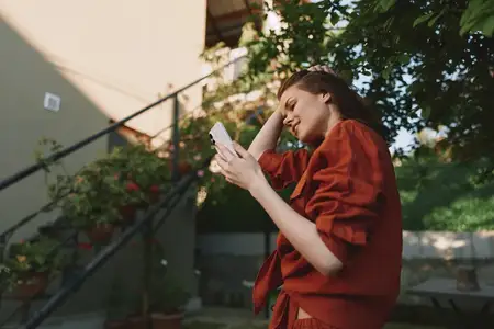 Сцена из жизни на открытом воздухе: сосредоточенная, жизнерадостная женщина с телефоном в теплой обстановке, окруженная зеленью, повседневная мода и расслабленная поза