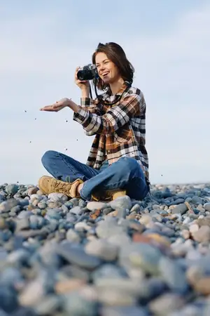 Повседневный фотограф у моря с фотоаппаратом, сидящий на камнях, наслаждающийся солнечным днем, природой вокруг, сосредоточенный на творчестве, хобби и создании воспоминаний у воды