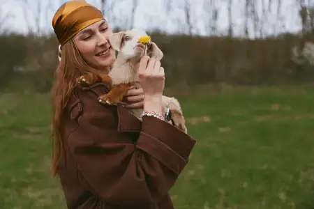 Молодая женщина в коричневом пальто и подходящем платке нежно держит козлёнка, улыбаясь и протягивая жёлтый цветок на зелёном поле с голыми деревьями на заднем плане