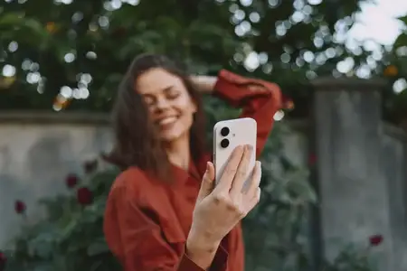 Улыбающаяся женщина в повседневной модной куртке делает уличное портретное селфи, празднуя моменты жизни с помощью смартфона, при естественном освещении и теплом садовом фоне