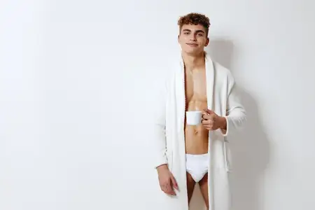 Молодой человек улыбается, наслаждаясь чашкой кофе, одетый в халат и нижнее белье, на минималистичном белом фоне