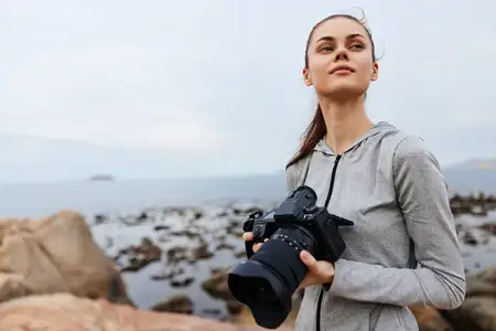 Уверенная молодая женщина-фотограф в серой толстовке держит камеру на каменистом пляже, запечатлевая суть творчества и приключений в облачный день