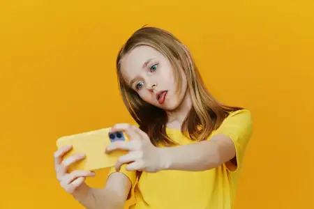 Веселая девушка с длинными волосами игриво делает селфи на смартфон, одетая в ярко-желтую футболку на сплошном желтом фоне, передавая юношескую энергию и креативность