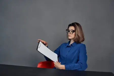 Женщина в синей рубашке сидит за столом, держа в руках папку с документами