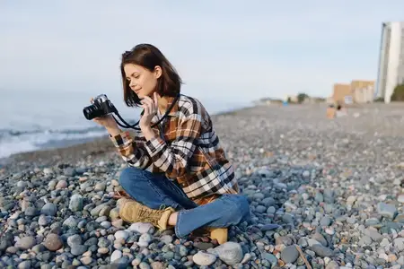 Фотограф в повседневной одежде на каменистом пляже: женщина присела с камерой, на ней клетчатая рубашка и ботинки