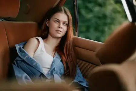 Молодая женщина с длинными каштановыми волосами отдыхает в машине, одетая в белую майку и синюю джинсовую куртку