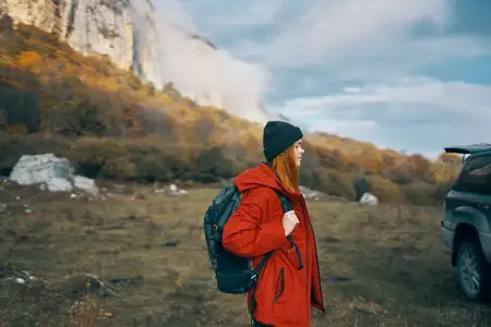 Женщина-модель в красной куртке с рюкзаком осенью на улице, в теплой шапке, среди опавших листьев