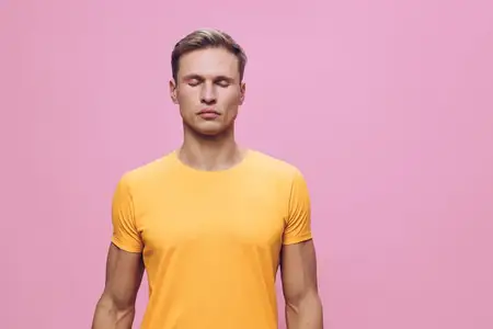 Молодой человек медитирует с закрытыми глазами, одетый в оранжевую футболку, стоит изолированно на розовом фоне, демонстрируя осознанность и благополучие ментального здоровья