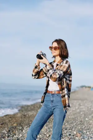 Женщина-фотограф на берегу с камерой; одета в клетчатую рубашку и джинсы; стоит на открытом воздухе вдоль побережья; кадрирует приморский момент с фокусом; объектив в поле зрения; естественный свет…