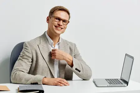 Счастливый молодой человек в очках и светло-сером пиджаке работает за ноутбуком за столом в чистом, минималистичном офисном помещении, уверенно улыбается, профессиональный вид, офисная работа…