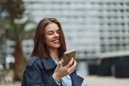 Весёлая молодая женщина в тёмном пальто держит смартфон на улице, улыбается и смотрит в сторону