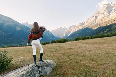 Вид со спины: женщина в коричневой куртке и ботинках стоит на камне посреди открытого луга, глядя на панораму гор и долины