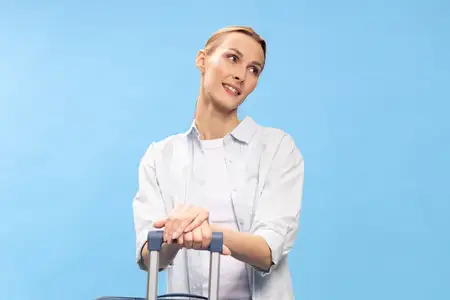Улыбающаяся молодая женщина держится за ручку чемодана, одетая в белую повседневную одежду, и смотрит в сторону на синем фоне