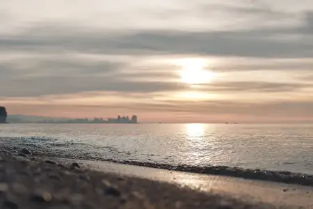 Спокойный берег моря на закате с мягким светом на песке, отражающей водой и далеким горизонтом города, запечатлевающий мирное вечернее настроение и бескрайний горизонт над морем