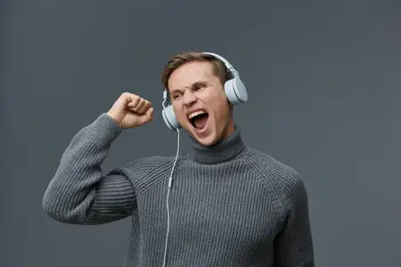 Счастливый молодой человек с радостным выражением лица, одетый в повседневный серый свитер и слушающий музыку в больших накладных наушниках, празднует или наслаждается моментом музыки и радости на…
