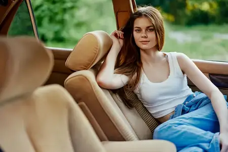 Расслабленная молодая женщина сидит в винтажном автомобиле, одетая повседневно, излучая спокойную и беззаботную атмосферу, а пышный зеленый фон усиливает безмятежное настроение