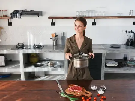 Женщина на кухне с кастрюлей в руках улыбается, готовит ужин из мяса и овощей на деревянном столе на фоне стильной кухни