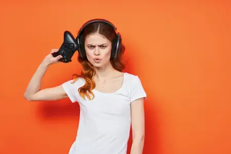 Женщина в белой футболке и наушниках играет с джойстиком на оранжевом фоне