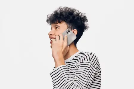 Молодой человек с вьющимися волосами разговаривает по смартфону, одетый в черно-белую полосатую рубашку с длинным рукавом, улыбаясь с расслабленным и веселым выражением лица на чистом белом фоне