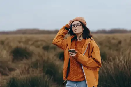 Женщина на улице в поле, в шапке и очках, оранжевая куртка поверх вязаного свитера