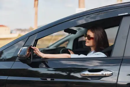Женщина на водительском сиденье автомобиля, положив руку на руль, задумчиво смотрит в окно, запечатлевая момент размышлений и путешествия