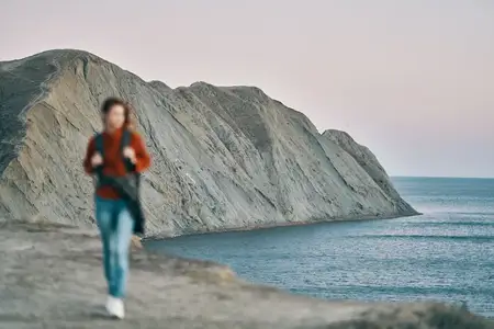 Женщина с рюкзаком, в свитере, на фоне гор и моря, наслаждается свежим воздухом природы