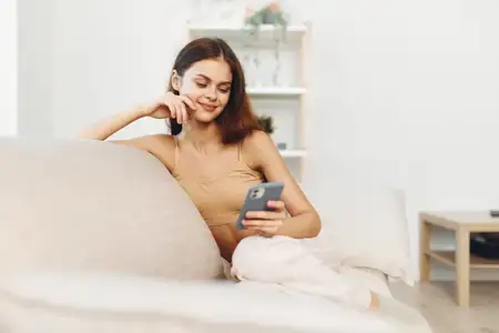 Улыбающаяся женщина держит мобильный телефон, отдыхая на уютном диване в современной гостиной