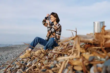Повседневный уличный фотограф на берегу: смотрит в объектив, документируя волны, свет и текстуру коряги в расслабленный творческий момент