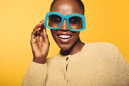 Радостная темнокожая африканская женщина позирует в больших синих очках и уютном желтом свитере на желтом фоне