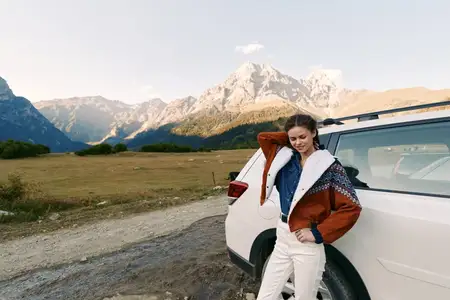 Женщина у машины на проселочной дороге с горами и широким пейзажем, одетая в уютную куртку для путешествия и приключений на природе с солнечным светом на альпийских вершинах
