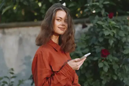 Естественный портрет женщины на открытом воздухе в лайфстайл-тематике, смартфон в руке, теплый свет, зеленый фон, откровенный повседневный момент в саду