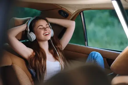 Молодая женщина с длинными волосами отдыхает в автомобиле, надев наушники и улыбаясь, слушая музыку, воплощая радость и безмятежность в солнечный день