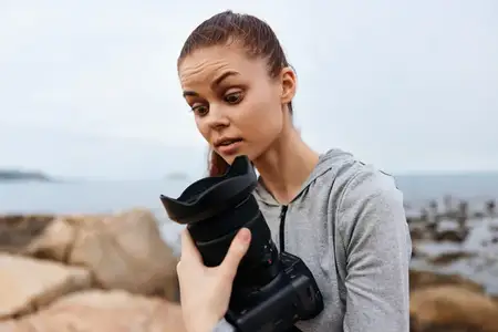 Удивленная молодая женщина в серой толстовке рассматривает камеру на каменистом пляже, передавая атмосферу побережья и эмоции любопытства и заинтригованности