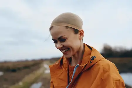 Улыбающаяся женщина на открытом воздухе, оранжевая куртка, шарф и шапка, спокойный портрет в профиль во время прогулки по сельской дороге, сельский пейзаж с холмами, облачное небо и мягкий свет