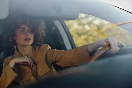 Женщина за рулём автомобиля в коричневой куртке, взгляд устремлён наружу
