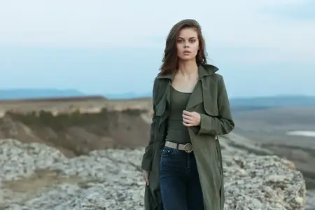 Спокойная женщина в плаще и джинсах стоит на скалистом холме, откуда открывается вид на величественные горы