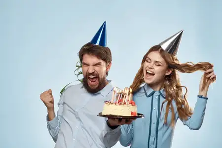 Мужчина и женщина празднуют день рождения с тортом и в праздничных колпаках на синем фоне