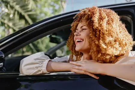 Улыбающаяся женщина с вьющимися волосами выглядывает из окна автомобиля, нежась в теплом солнечном свете со счастливым выражением лица, в окружении зелени и современной архитектуры