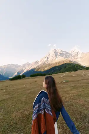 Женщина, закутанная в узорчатую шаль, на травянистом лугу смотрит в сторону далеких гор