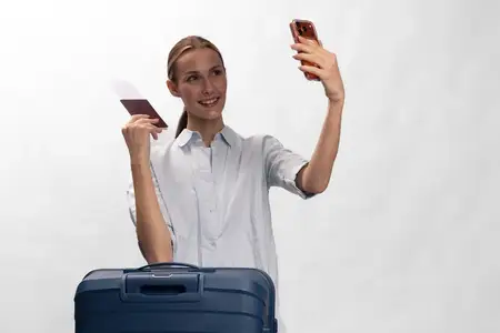 Улыбающаяся молодая женщина с чемоданом держит паспорт и мобильный телефон, делая селфи на белом фоне