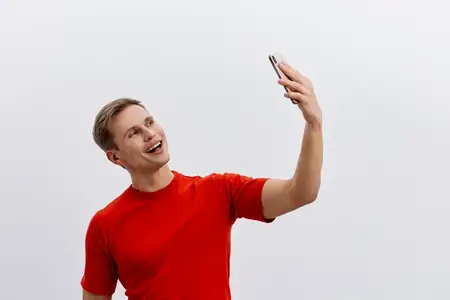 Улыбающийся мужчина в красной футболке держит смартфон и участвует в видеозвонке, изолированный на белом фоне