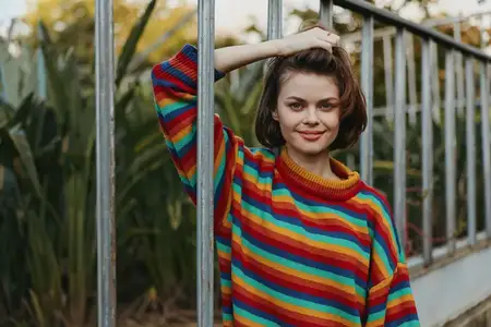 Портрет женщины в разноцветном свитере, улыбающейся и опирающейся на металлический забор на открытом воздухе