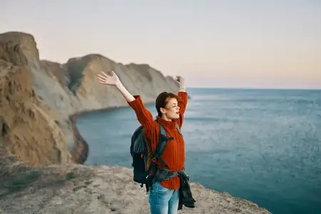 Счастливая путешественница в горах у моря жестикулирует руками, за спиной у неё рюкзак