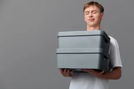 Молодой работник мужского пола демонстрирует стопки серых коробок для доставки и хранения, подчёркивая упаковку, управление запасами и складское хозяйство в минималистичной студийной сцене