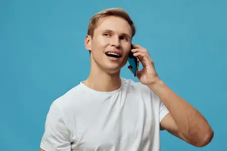 Улыбающийся мужчина разговаривает по телефону в повседневной белой футболке на синем фоне, выражая счастье и общение, что представляет образ жизни цифрового кочевника и связь через мобильные…