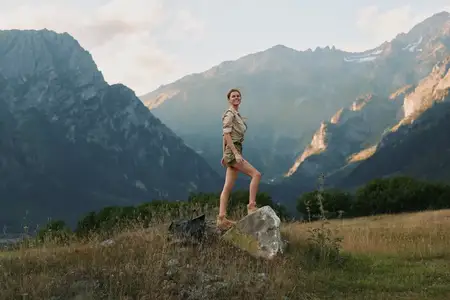 Молодая женщина в бежевой рубашке и шортах позирует на большом камне на травянистом горном лугу, на заднем плане возвышаются величественные пики под частично облачным небом