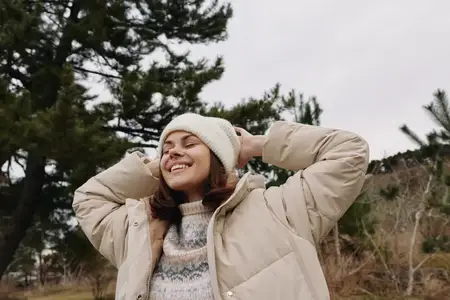 Улыбающаяся женщина в вязаном свитере и куртке стоит в естественной обстановке, излучая комфорт и радость во время сегодняшней съёмки на свежем воздухе