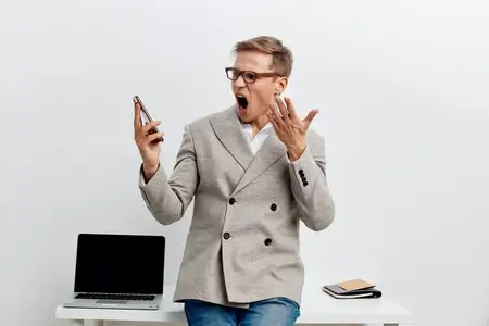 Удивленный мужчина в очках держит смартфон, выглядит потрясенным, одет в клетчатую куртку, сидит за столом с ноутбуком на белом фоне